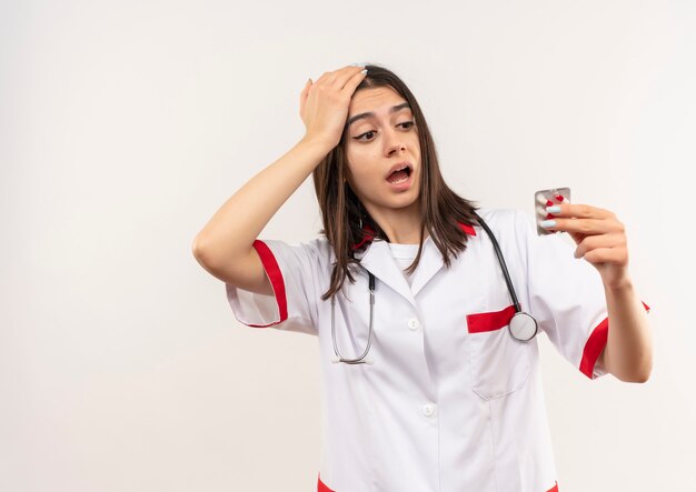 Giovane medico femminile in camice bianco con lo stetoscopio intorno al collo che tiene la bolla con le pillole guardandolo confuso in piedi sopra il muro bianco