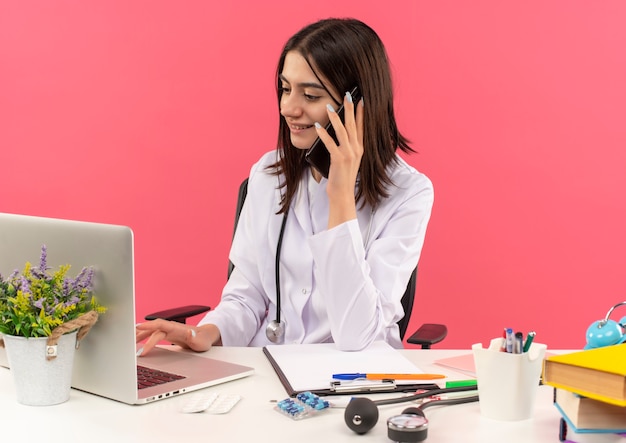 Giovane medico femminile in camice bianco con lo stetoscopio intorno al collo lavorando sul computer portatile e parlando al telefono cellulare con il sorriso sul viso seduto al tavolo sopra il muro rosa