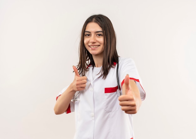 Giovane medico femminile in camice bianco con lo stetoscopio intorno al collo sorridendo allegramente mostrando i pollici in su in piedi sul muro bianco