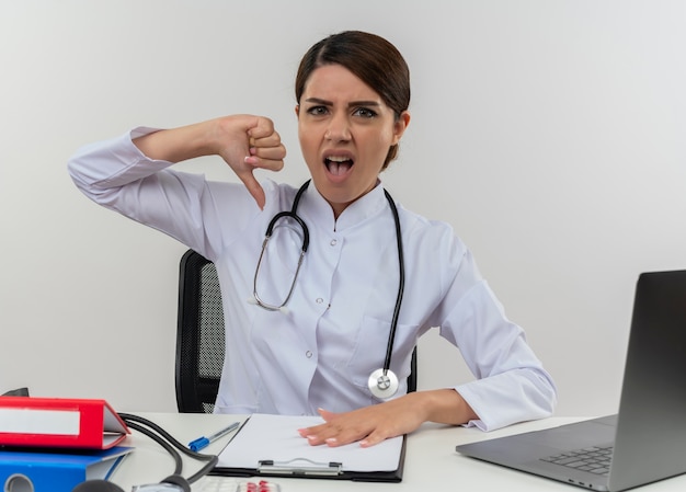 Giovane medico femminile infastidito che indossa veste medica e stetoscopio seduto alla scrivania con strumenti medici e laptop che mostra il pollice verso il basso isolato sul muro bianco