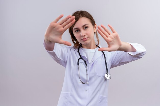 Giovane medico femminile infelice che porta veste medica e stetoscopio che fa gesto di arresto sulla parete bianca isolata con lo spazio della copia