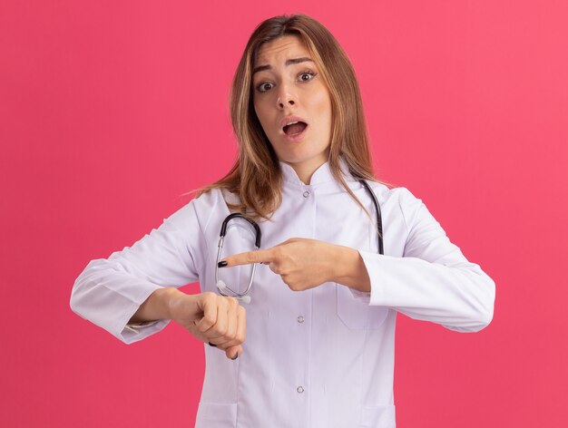 Giovane medico femminile preoccupato che indossa veste medica con lo stetoscopio che mostra il gesto dell'orologio da polso isolato sulla parete rosa