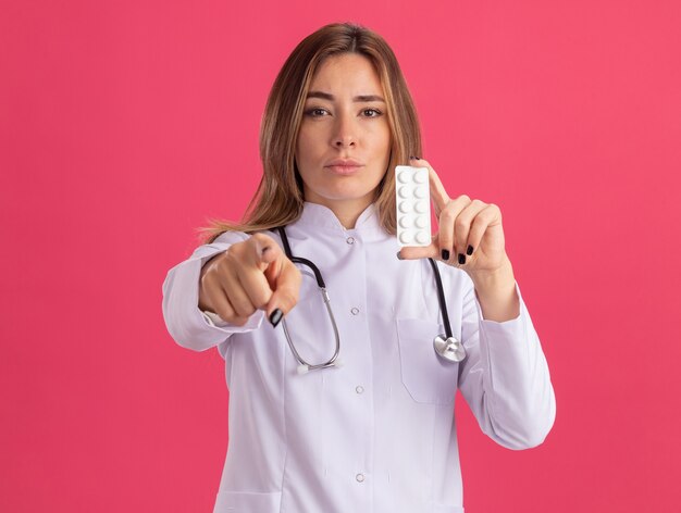 Giovane medico femminile sicuro che porta veste medica con le pillole della tenuta dello stetoscopio che vi mostrano gesto isolato sulla parete rosa