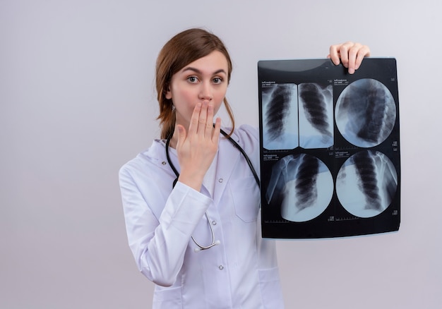 Giovane medico femminile sorpreso che indossa veste medica e stetoscopio e che tiene i raggi x ha sparato sulla parete bianca isolata con lo spazio della copia