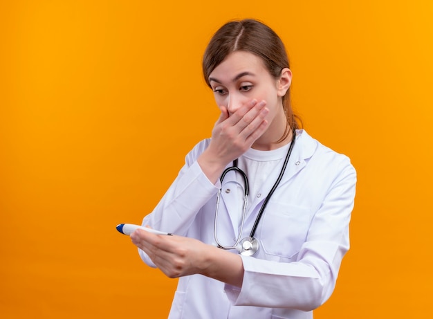 Giovane medico femminile sorpreso che indossa veste medica e stetoscopio e che tiene il termometro guardandolo con la mano sulla bocca sulla parete arancione isolata con lo spazio della copia
