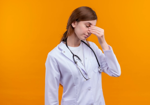 Giovane medico femminile stanco che indossa veste medica e stetoscopio mettendo la mano sul naso sulla parete arancione isolata con lo spazio della copia