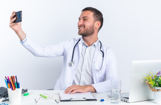 Giovane medico in camice bianco e con lo stetoscopio facendo selfie utilizzando smartphone felice e positivo sorridente allegramente seduto al tavolo con il computer portatile su bianco