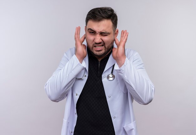 Giovane medico maschio barbuto che indossa camice bianco con stetoscopio che chiude le orecchie con le dita con espressione infastidita per il rumore del suono forte