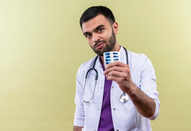 giovane medico maschio che indossa abito medico e stetoscopio tenendo le pillole sul muro viola isolato