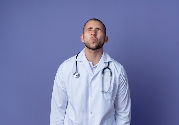 Giovane medico maschio che indossa veste medica e stetoscopio intorno al collo facendo gesto di bacio con gli occhi chiusi isolato su sfondo viola con spazio di copia