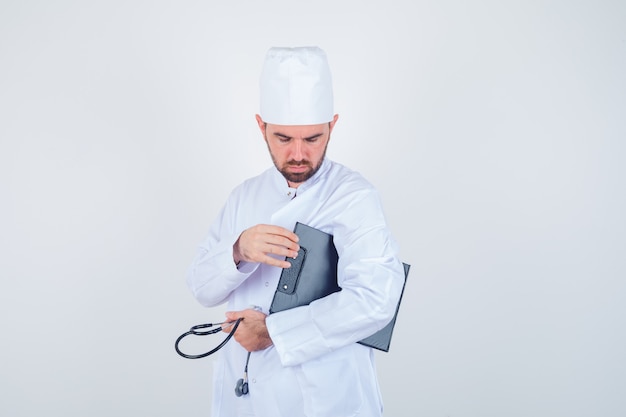Giovane medico maschio che tiene appunti e stetoscopio in uniforme bianca e guardando premuroso, vista frontale.