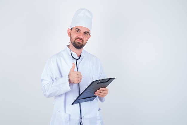 Giovane medico maschio che tiene appunti, mostrando il pollice in su in uniforme bianca e guardando fiducioso. vista frontale.