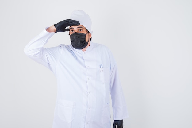 Giovane medico maschio che tiene la mano sulla fronte mentre distoglie lo sguardo in uniforme bianca e sembra concentrato. vista frontale.