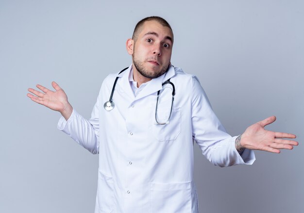 Giovane medico maschio confuso che porta veste medica e stetoscopio intorno al suo collo che mostra le mani vuote isolate su bianco