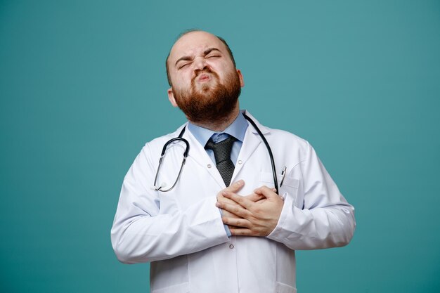 Giovane medico maschio dolorante che indossa camice medico e stetoscopio intorno al collo tenendo le mani sul cuore con angoscia con gli occhi chiusi isolati su sfondo blu
