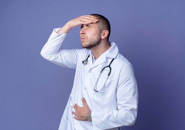 Giovane medico maschio dolorante che indossa veste medica e stetoscopio che mette la mano sulla testa e sulla pancia con gli occhi chiusi isolati sulla porpora con lo spazio della copia