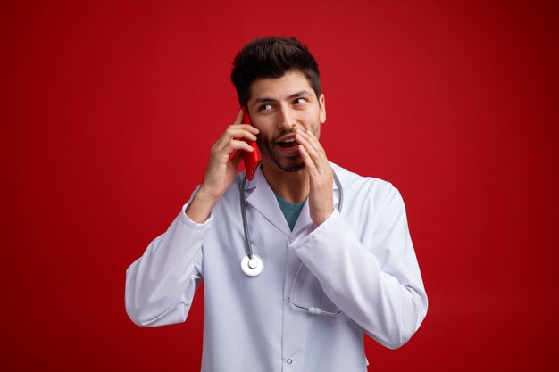 Giovane medico maschio gioioso che indossa uniforme medica e stetoscopio intorno al collo tenendo la mano vicino alla bocca parlando al telefono guardando il sussurro laterale isolato su sfondo rosso