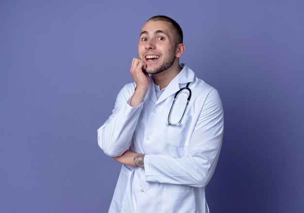 Giovane medico maschio impressionato che porta veste medica e stetoscopio che mette le mani sul mento e sotto il gomito isolato sulla porpora