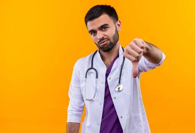 giovane medico maschio indossa abito medico stetoscopio il pollice verso il basso sulla parete gialla isolata