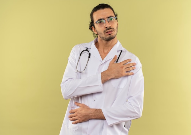 Giovane medico maschio malato con vetri ottici che indossa una veste bianca con lo stetoscopio che mette la mano sulla spalla dolorante sul verde