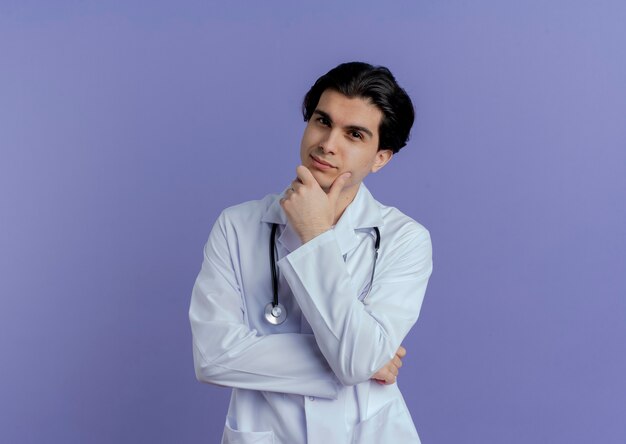 Giovane medico maschio premuroso che indossa veste medica e stetoscopio che tocca il mento isolato sulla parete viola con lo spazio della copia