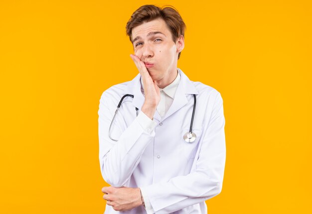 Giovane medico maschio scontento che indossa una tunica medica con uno stetoscopio che mette la mano sulla guancia