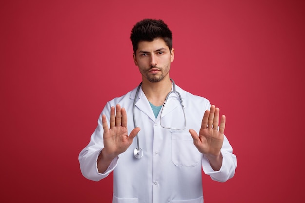 Giovane medico maschio serio che indossa uniforme medica e stetoscopio intorno al collo guardando la fotocamera che mostra il gesto di arresto con entrambe le mani isolate su sfondo rosso