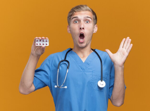 Giovane medico maschio sorpreso che indossa l'uniforme del medico con lo stetoscopio che tiene le pillole che diffondono la mano isolata sulla parete arancio