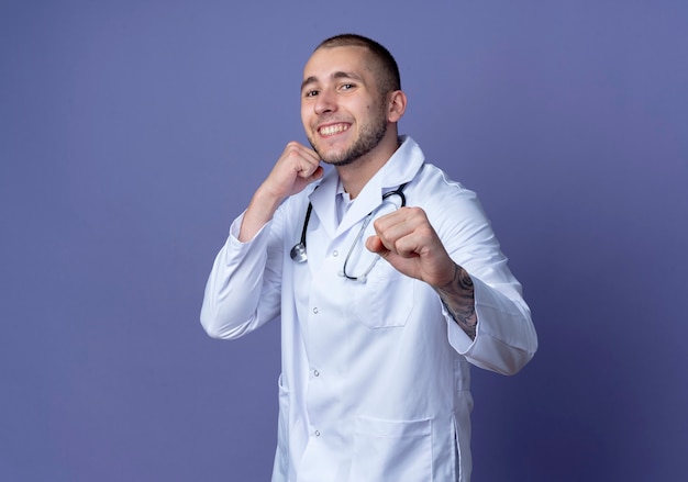 Giovane medico maschio sorridente che indossa veste medica e stetoscopio che fa gesto di boxe nella parte anteriore isolata sulla parete viola