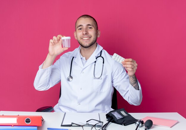 Giovane medico maschio sorridente che indossa veste medica e stetoscopio seduto alla scrivania con strumenti di lavoro che tengono becher medico e confezione di compresse isolato sulla parete rosa