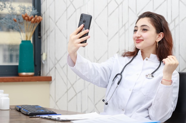Giovane medico seduto in ufficio e prendendo selife Foto di alta qualità