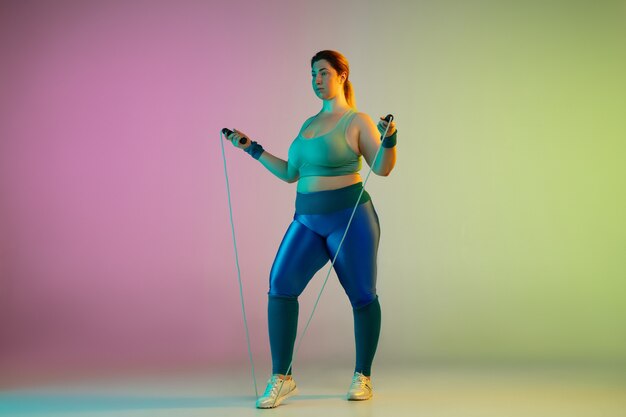 Giovane modello femminile caucasico plus size allenamento sulla parete verde viola sfumata al neon. Fare esercizi di allenamento con la corda per saltare.