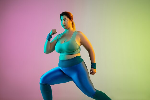 Giovane modello femminile caucasico plus size allenamento sulla parete verde viola sfumata al neon. Fare esercizi di stretching.