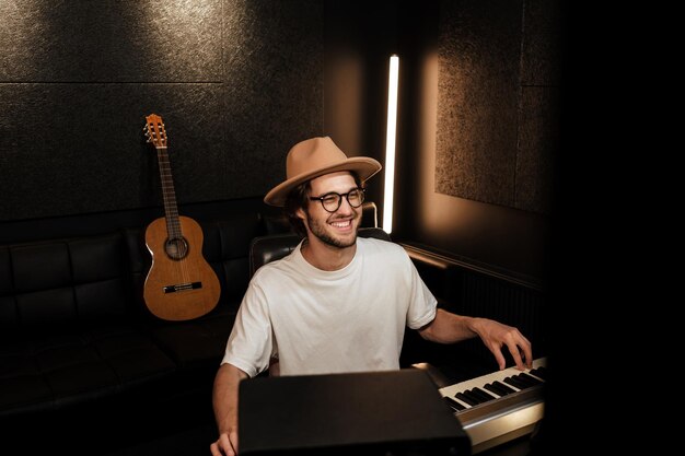 Giovane musicista bello in cappello che compone felicemente la nuova canzone nel moderno studio di registrazione del suono