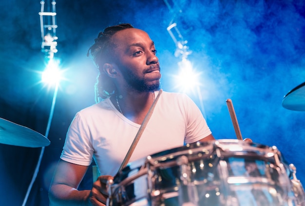 Giovane musicista jazz afro-americano o batterista che suona la batteria su sfondo blu in fumo incandescente intorno a lui.