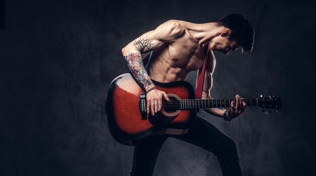 Giovane musicista senza camicia bello che suona la chitarra mentre salta. Isolato su uno sfondo scuro.