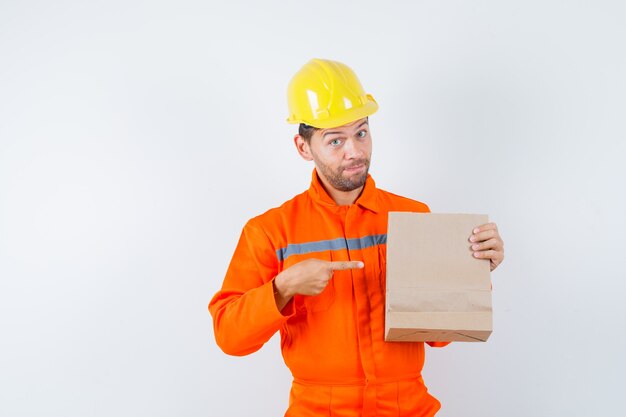 Giovane operaio che indica al sacco di carta in uniforme, casco.