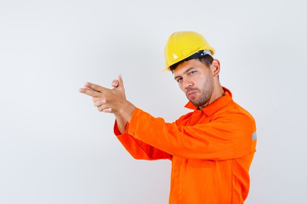 Giovane operaio che mostra gesto di tiro in uniforme, casco e guardando fiducioso.