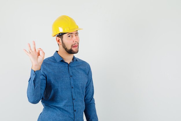 Giovane operaio che mostra gesto giusto, occhi ammiccanti, che attacca fuori la lingua in camicia, casco