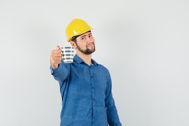 Giovane operaio che offre una tazza di caffè in camicia, casco e dall'aspetto gentile