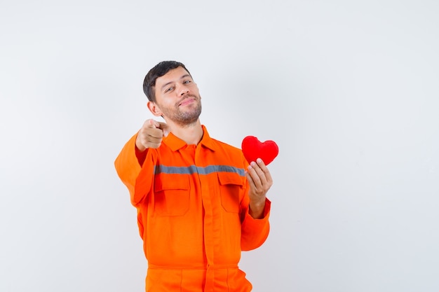 Giovane operaio che tiene cuore rosso, che indica davanti in uniforme e che sembra allegro.