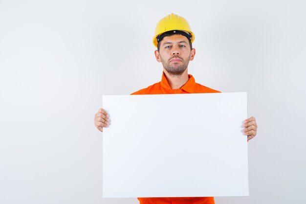 Giovane operaio che tiene tela bianca in uniforme, casco e che sembra fiducioso.