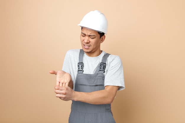 Giovane operaio edile dolorante che indossa il casco di sicurezza e uniforme che tiene la mano con gli occhi chiusi