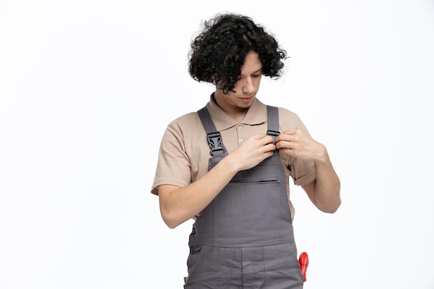 Giovane operaio edile maschio concentrato che indossa l'uniforme che fissa la sua uniforme con strumenti da costruzione in tasca isolato su sfondo bianco con spazio per la copia