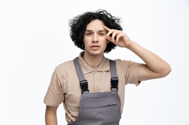 Giovane operaio edile maschio confuso che indossa la testa commovente uniforme che esamina la macchina fotografica isolata su fondo bianco