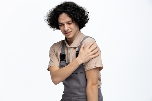 Giovane operaio edile maschio dolorante che indossa l'uniforme guardando in basso tenendo la mano sulla spalla isolata su sfondo bianco