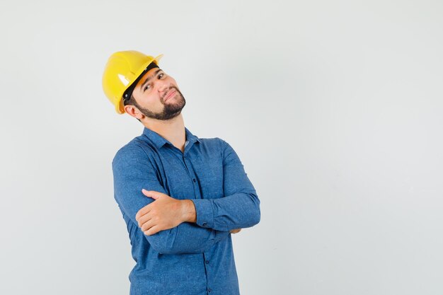 Giovane operaio in camicia, casco in piedi con le braccia incrociate e guardando allegro