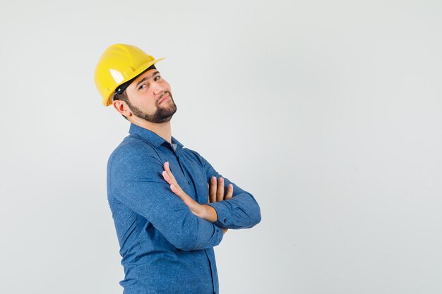 Giovane operaio in piedi con le braccia incrociate in camicia, casco e guardando fiducioso.