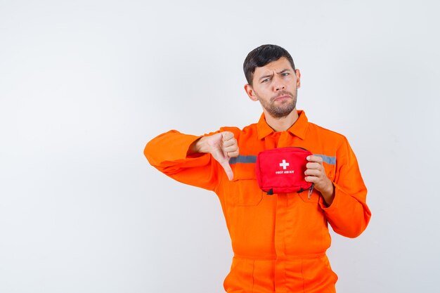 Giovane operaio in uniforme che tiene il kit di pronto soccorso, mostrando il pollice verso il basso e guardando insoddisfatto.