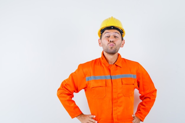 Giovane operaio mantenendo le labbra piegate in uniforme, casco e guardando allegro.